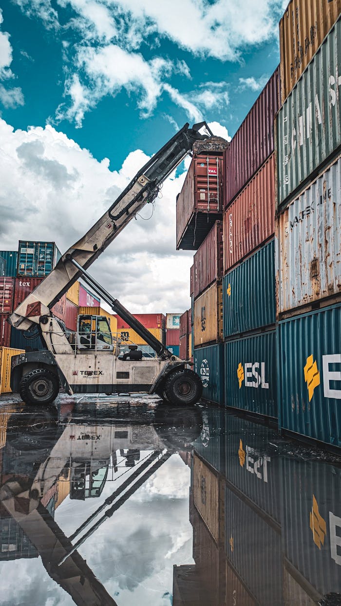 A crane loader stacking colorful shipping containers at a port with reflection in water.